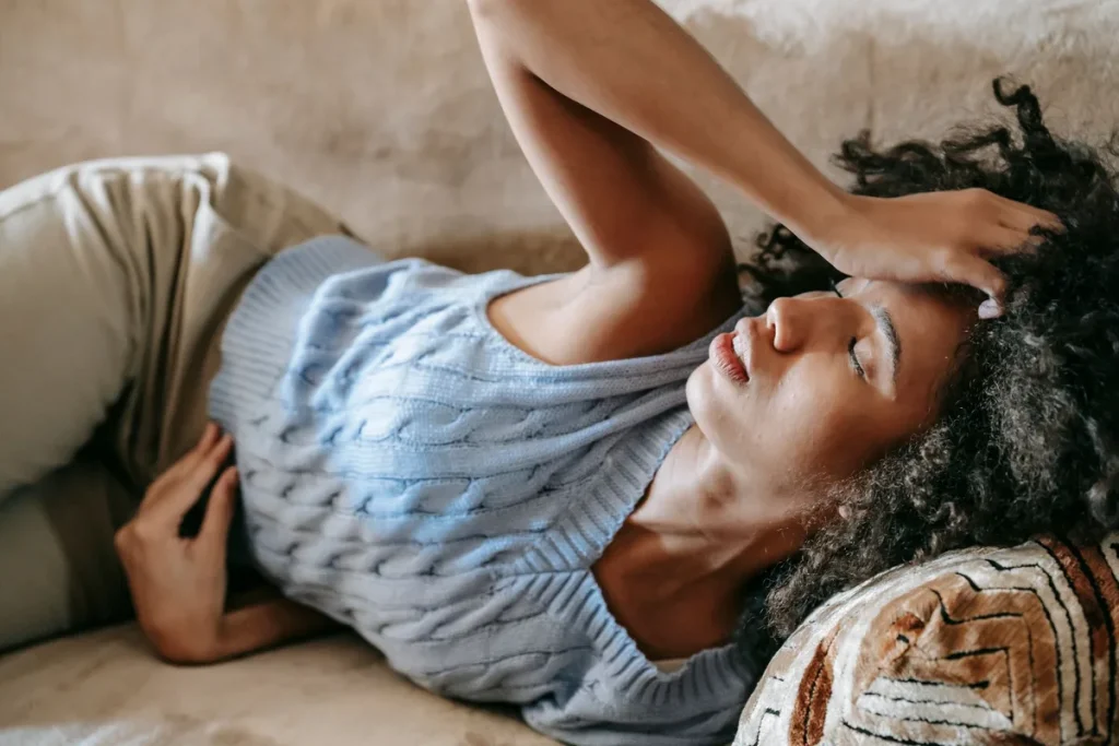 Femme en vêtements d'été assise sur un lit, main sur l'abdomen, expression inconfortable